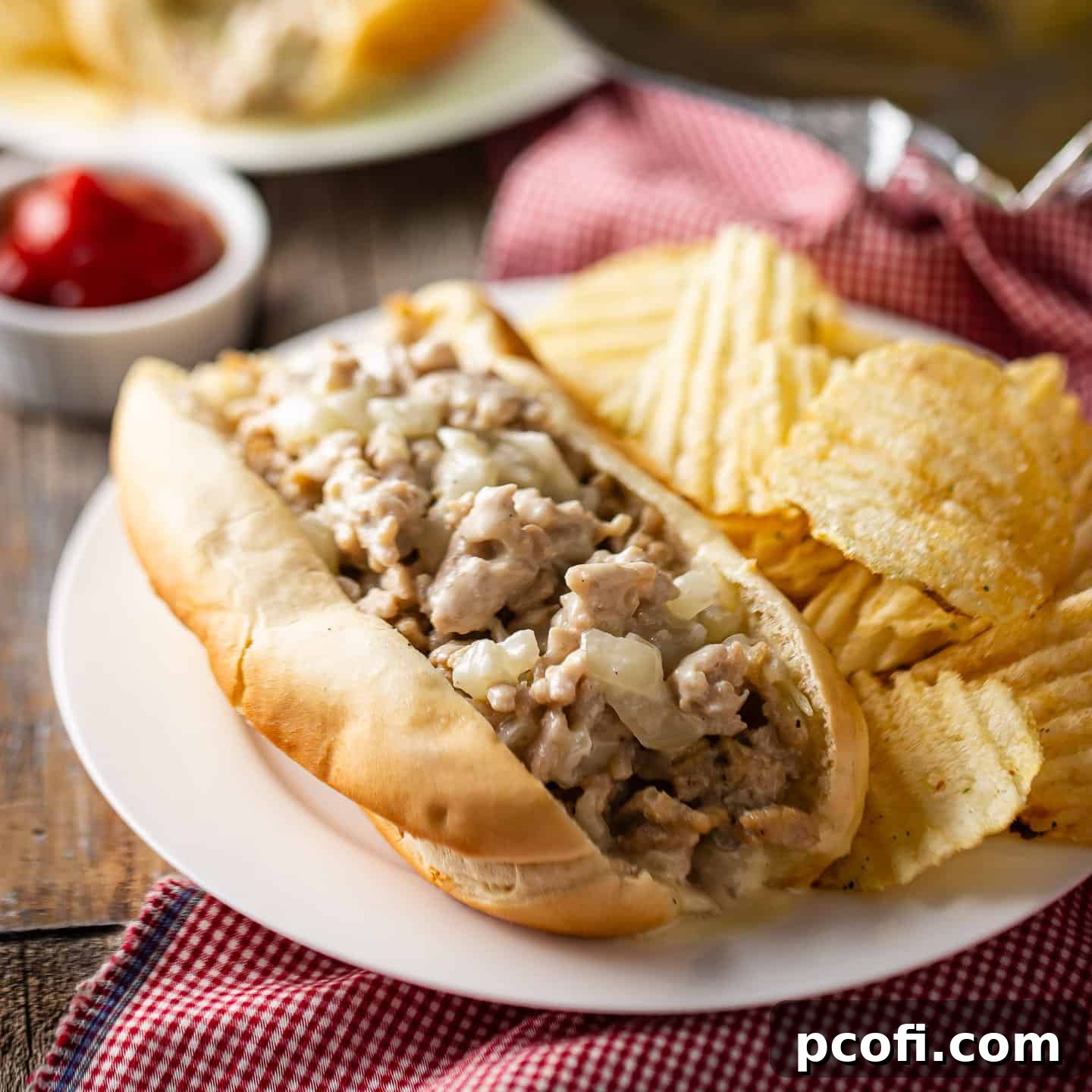 Homemade chicken cheesesteak on a white plate with potato chips and ketchup.