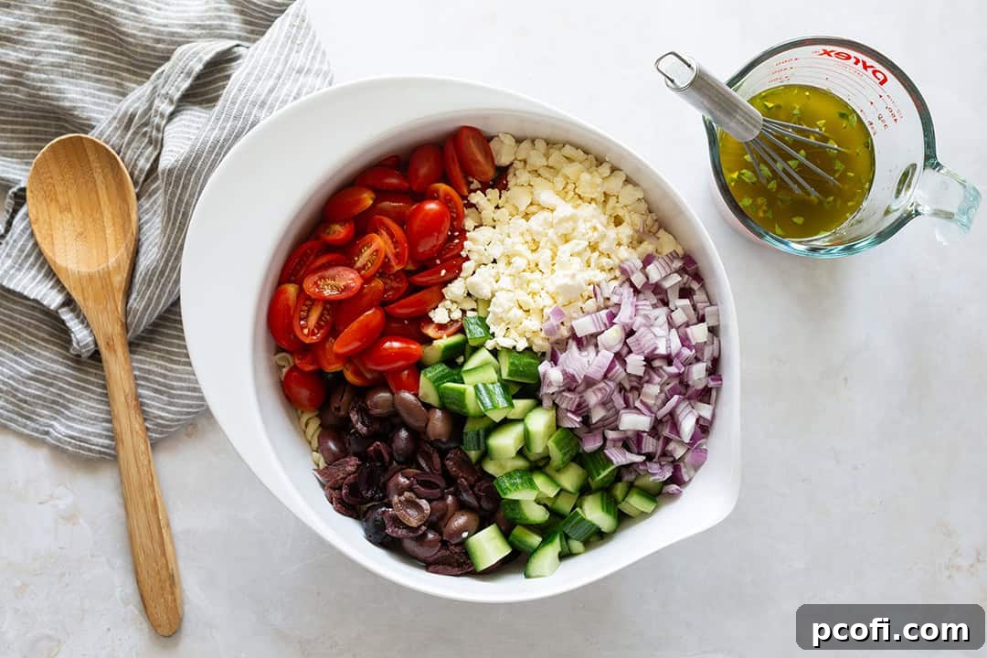 Ingredients for orzo pasta Greek salad in a bowl next to a measuring cup with vinaigrette, ready for mixing.