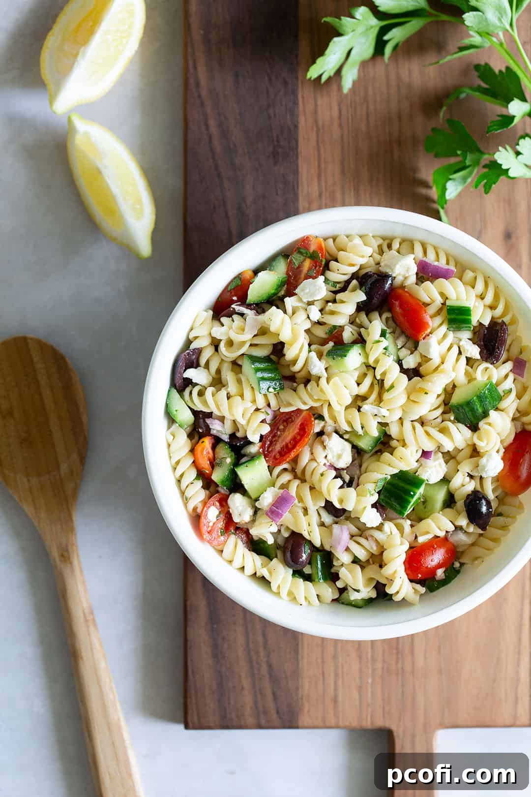 A beautiful recipe for Greek pasta salad in a bowl set on a rustic cutting board, garnished with fresh herbs.
