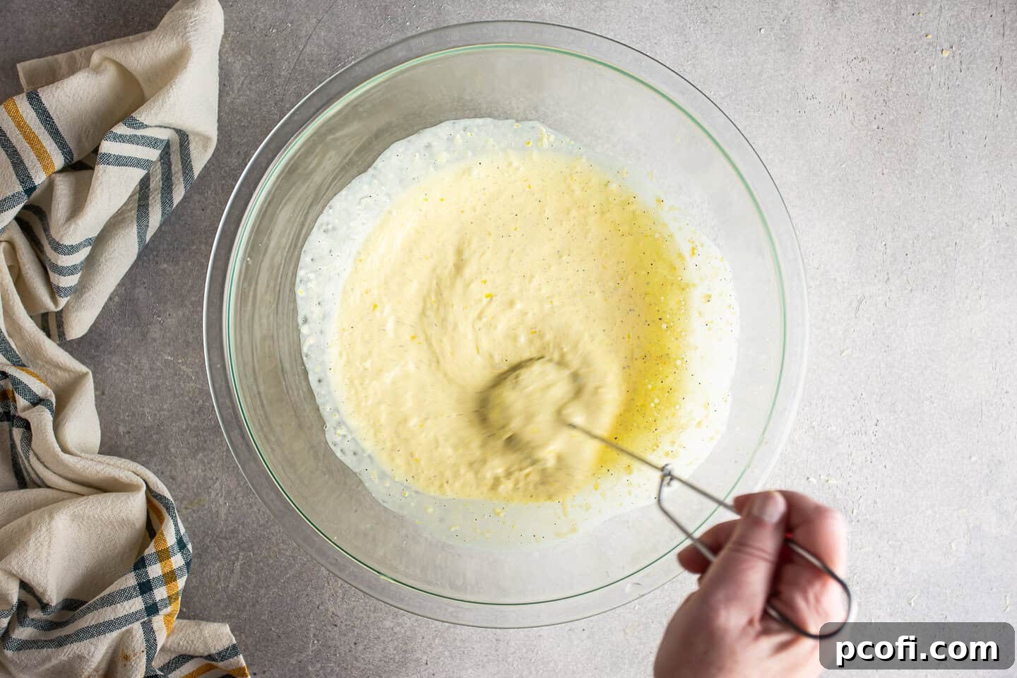 A hand is actively whisking together the creamy lemon pasta sauce ingredients in a large mixing bowl, illustrating the smooth, blended consistency being achieved. The mixture is light yellow and free of lumps, indicating a well-prepared sauce base.