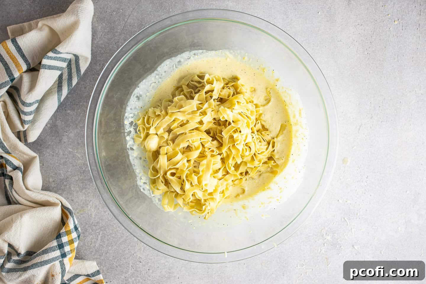 The process of adding cooked and buttered pasta into the prepared creamy lemon sauce in a large mixing bowl, illustrating the next stage of combining ingredients to create the finished dish. Steam rises gently from the hot pasta.
