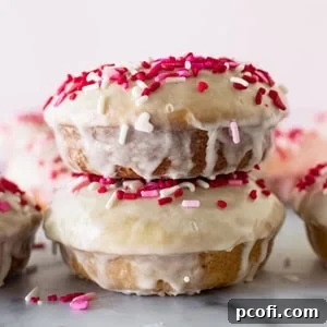 Stacked baked glazed donuts with festive sprinkles on a white surface.