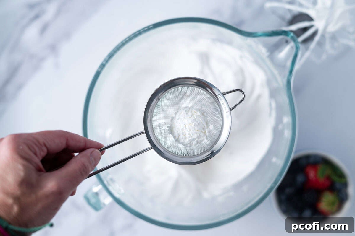 hand holding a sifter of cornstarch over a mixing bowl of meringue