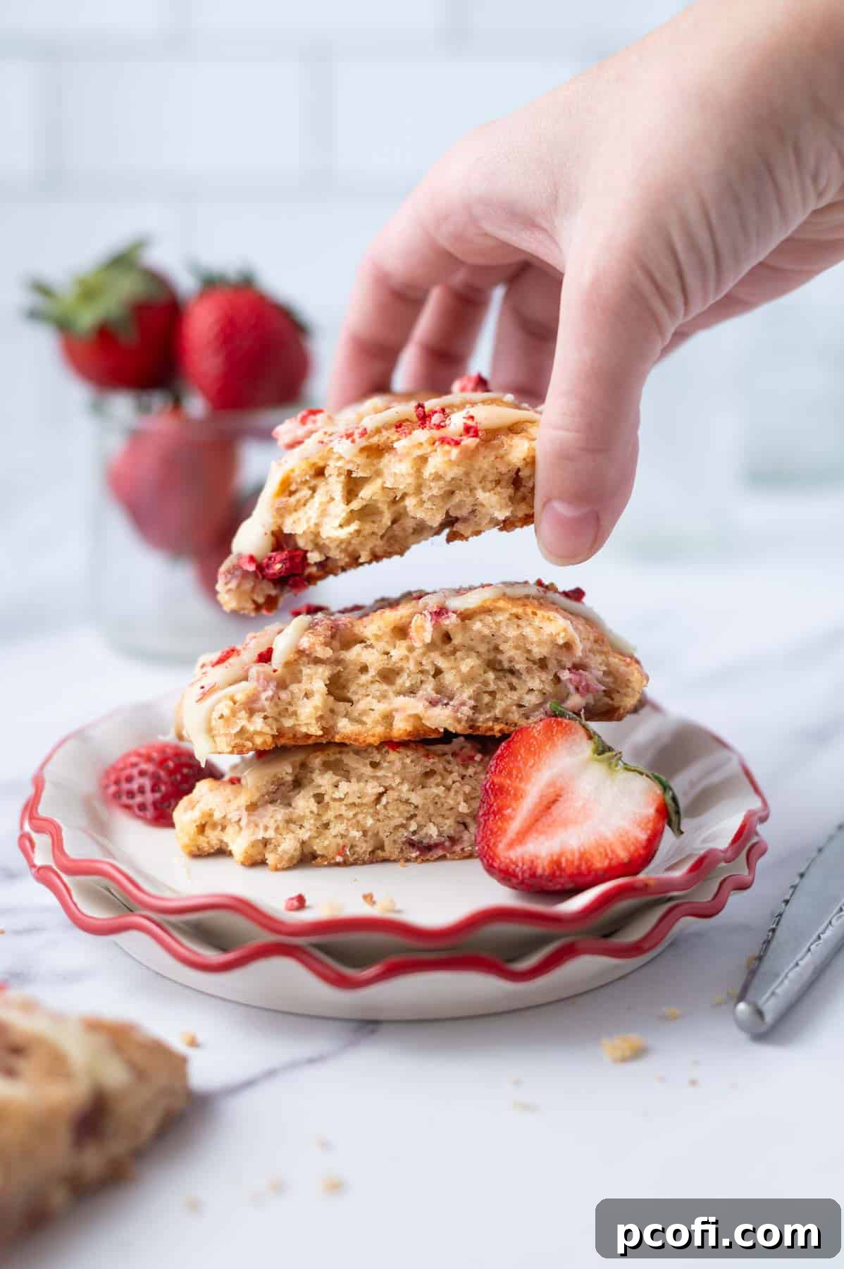 A hand reaching for a freshly baked strawberry scone from a plate, beautifully glazed and ready to enjoy.