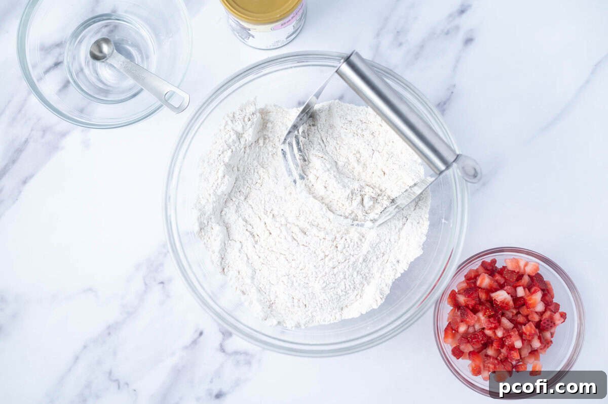A bowl filled with flour and other dry ingredients, alongside a pastry cutter and another bowl of chopped fresh strawberries.