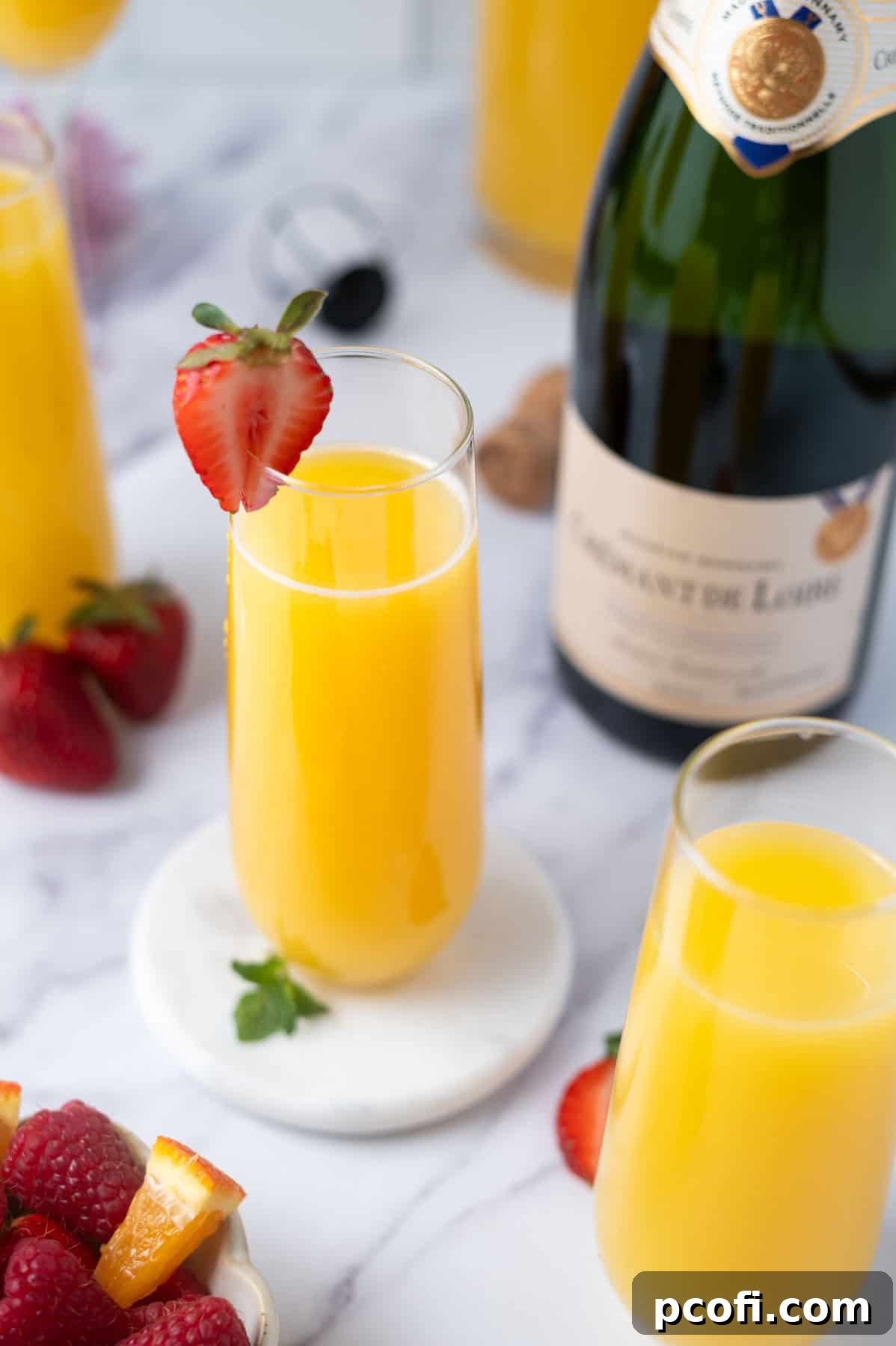 An array of mimosas in different glasses on a counter, with a bottle of sparkling wine and fresh fruit nearby.
