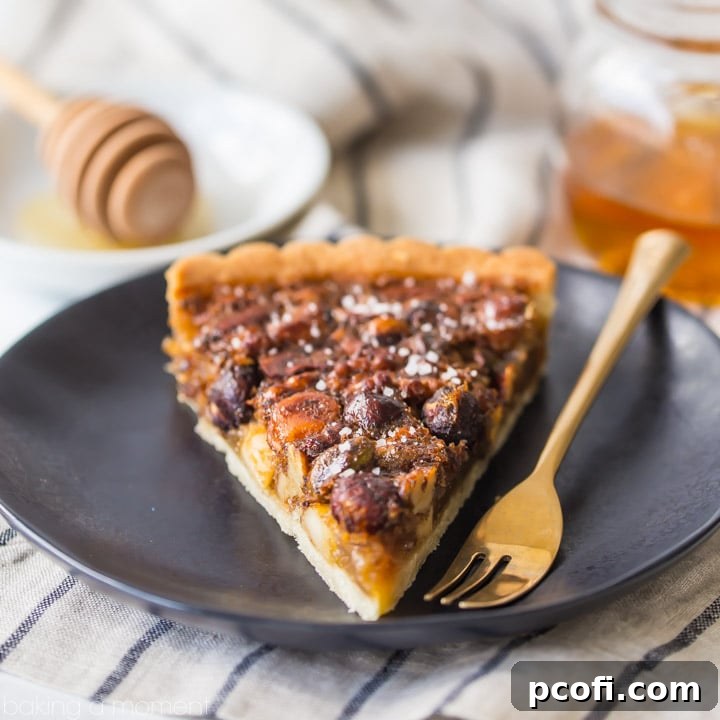 A slice of honey nut tart on a dark plate with gold fork and jar of honey in background