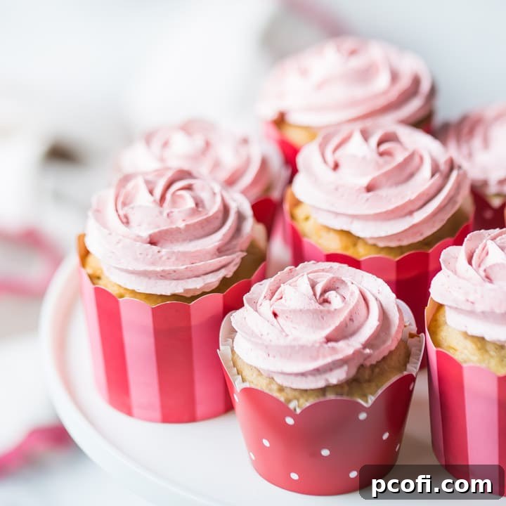 Strawberry cupcakes with strawberry buttercream, in pink, red, and white cupcake papers, sitting on a white cake stand. The buttercream has been piped in the shape of a rosette, and there is a pink-trimmed cloth in the background.