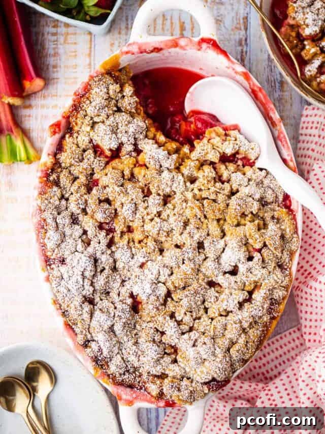 Overhead image of strawberry rhubarb crisp recipe, prepared and baked in an oval dish and surrounded with fresh strawberries and rhubarb stalks.