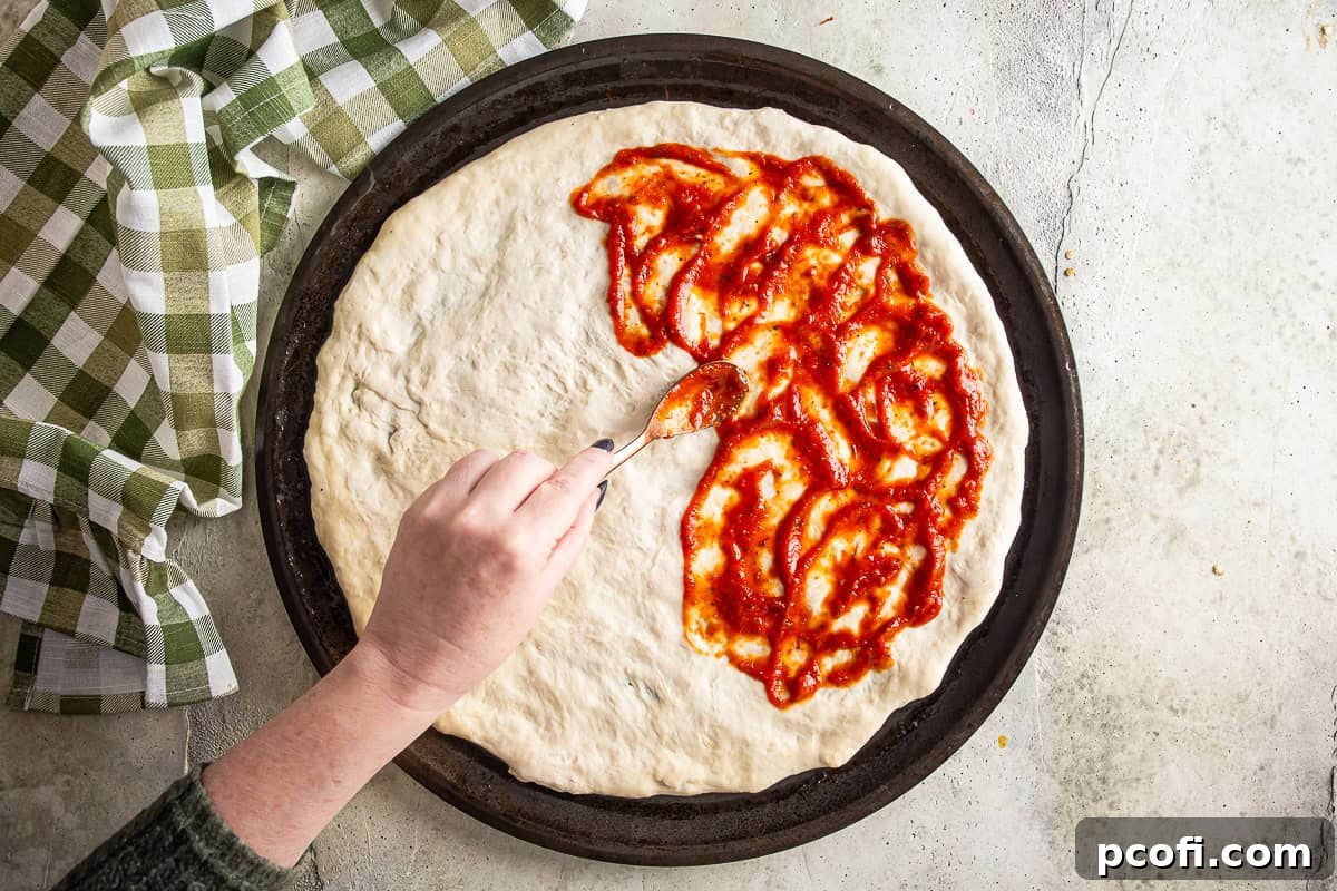 Adding a thin layer of pizza sauce onto stretched dough for jalapeno popper pizza.