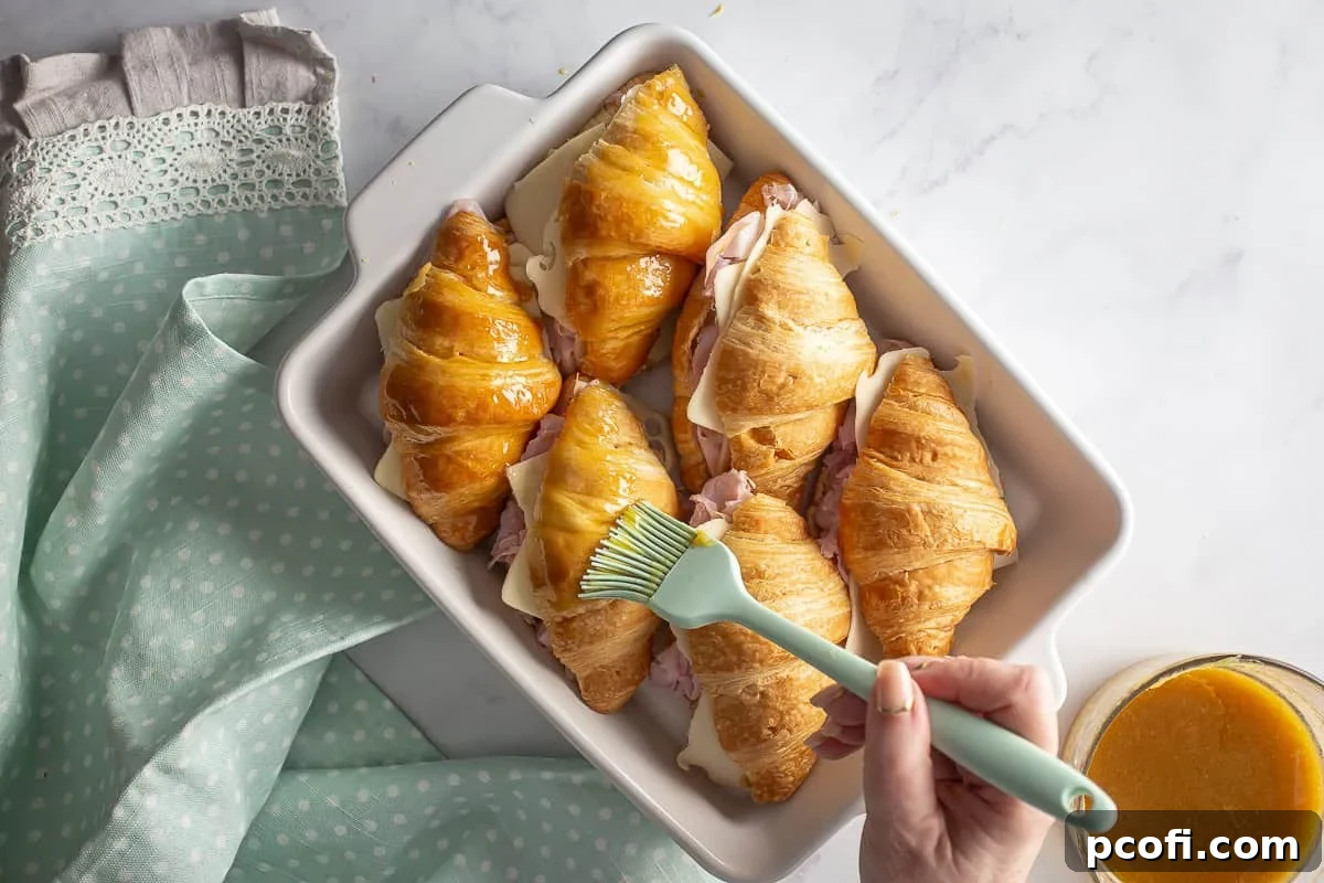 A hand brushing the Dijon-honey glaze generously over the tops of assembled ham and cheese croissant sandwiches.