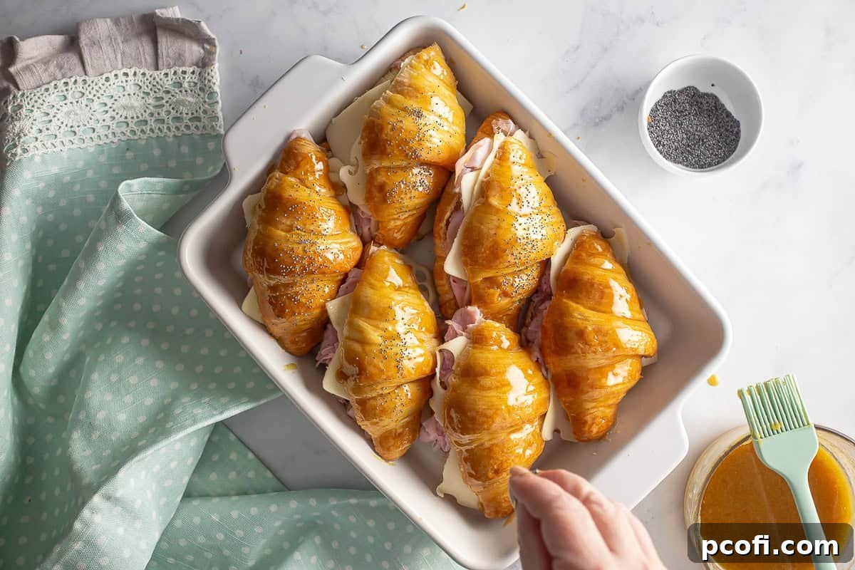 Poppy seeds being sprinkled liberally over the glazed tops of the ham and cheese croissant sandwiches, adding texture and visual appeal.