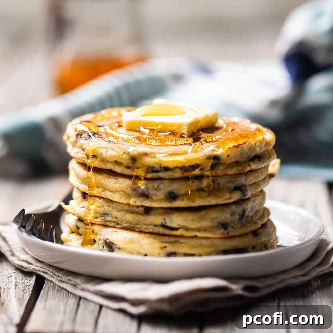 A stack of fluffy chocolate chip pancakes topped with butter and syrup on a white plate.