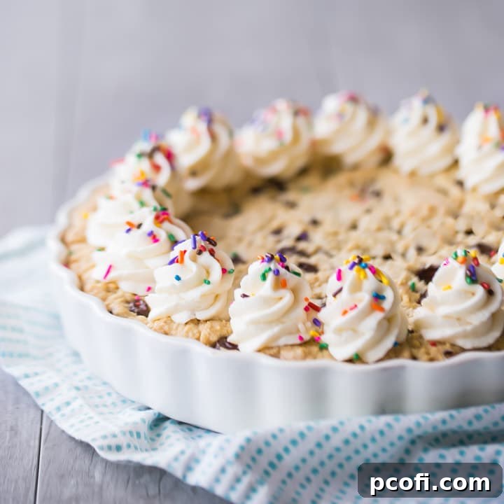 Square image of a giant chocolate chip cookie cake with vanilla buttercream rosettes and rainbow sprinkles on a turquoise cloth.
