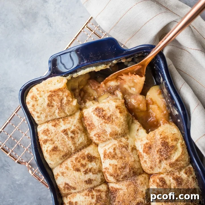 Snickerdoodle Apple Cobbler in a dark blue casserole dish: apple pie filling topped with cinnamon sugar snickerdoodle cookies, on a gray background with a copper serving spoon.